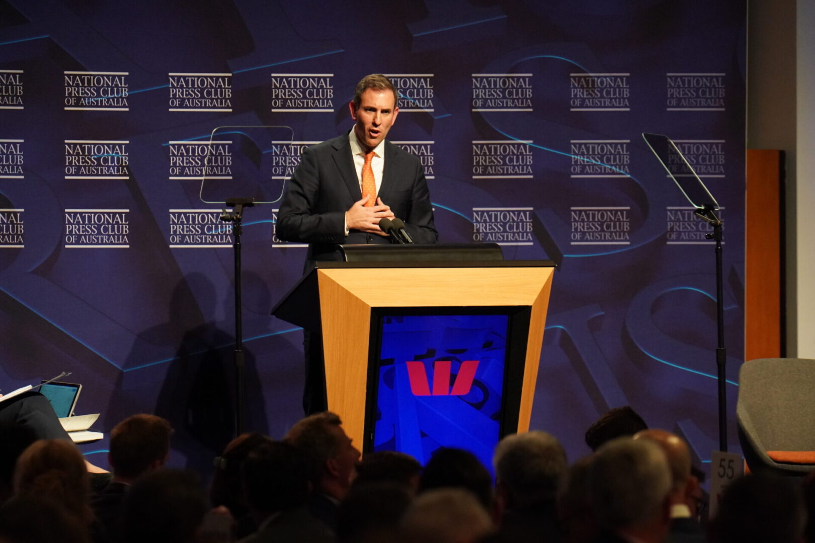 A man in suit and tie at podium with crowd.